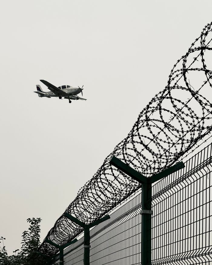 Small airplane flying past a barbed wire fence against a cloudy sky, suggesting travel and security.