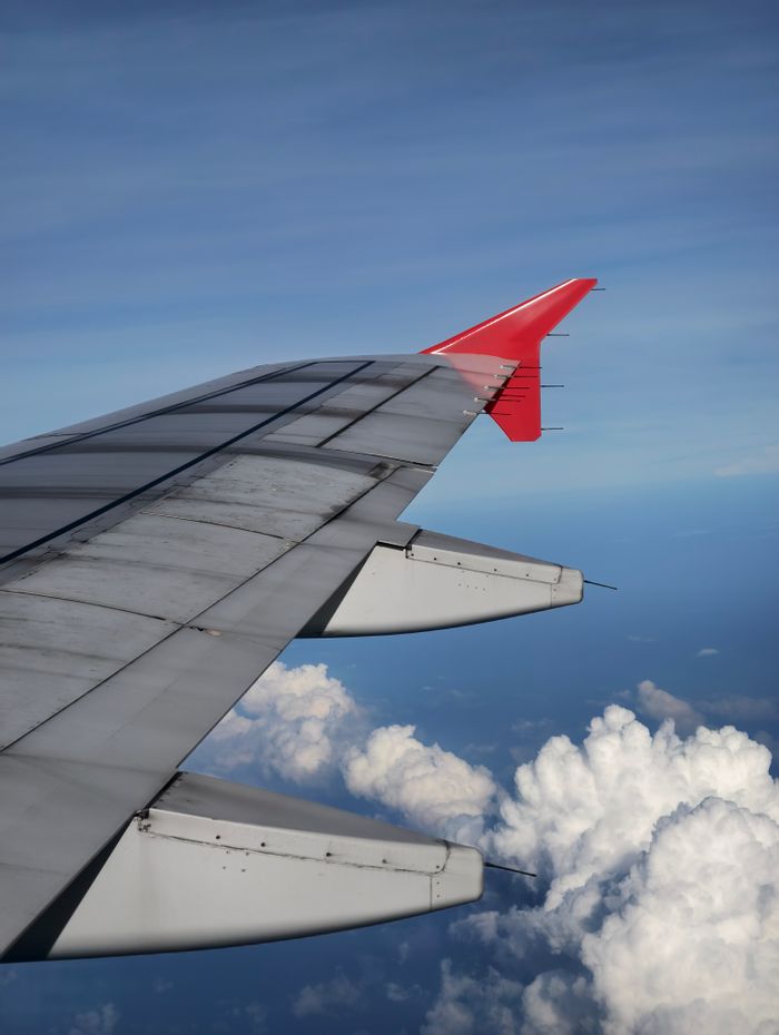 View of an airplane wing above fluffy white clouds and clear blue sky.